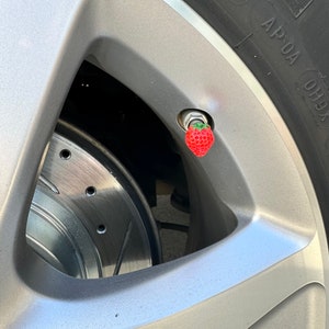 May include: Close-up of a red strawberry-shaped tire valve stem cap on a silver car wheel.