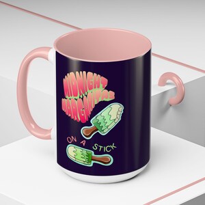 May include: A ceramic mug with a light pink interior and handle, featuring a dark purple exterior. The design includes the text "MIDNIGHT MARGARITAS ON A STICK" in pink and green, accompanied by two illustrations of green popsicles on sticks.