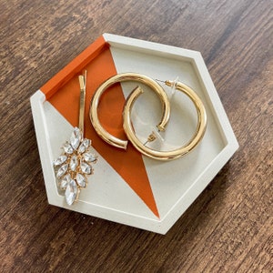 May include: A white and orange hexagonal tray with a geometric design. The tray is filled with gold hoop earrings and a crystal flower brooch.