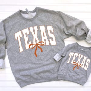 May include: Two grey sweatshirts with the word "TEXAS" in white letters and a brown bow. The larger sweatshirt has the word "TEXAS" in a larger font.