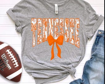 Könnte beinhalten: Ein graues T-Shirt mit dem Wort "TENNESSEE" in orangefarbenen, verwitterten Buchstaben und einer orangefarbenen Schleife. Ein brauner und weißer Football und blaue Jeans sind ebenfalls zu sehen.