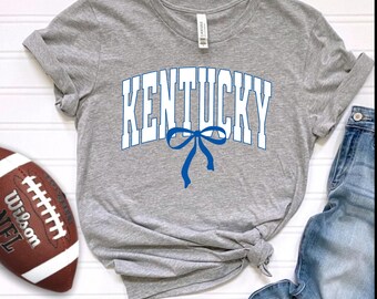 May include: A gray t-shirt with the word "KENTUCKY" in white block letters and a blue bow graphic. A brown football with white stitching and the word "Wilson" is on the left. Blue jeans are on the right.