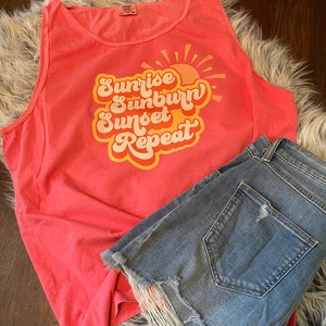 May include: A coral pink tank top with a graphic design that says "Sunrise Sunburn Sunset Repeat" in a retro font with a yellow sun graphic.