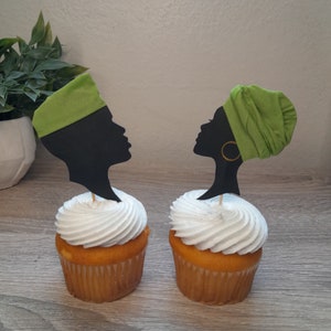 May include: Two cupcakes with white frosting topped with black silhouette cupcake toppers of a couple wearing traditional African headwear. The headwear is green.