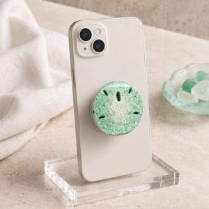 May include: A light-colored smartphone with a green and white pop-up phone grip. The grip has a glittery, speckled design with black accents. The phone is displayed on a clear acrylic stand.