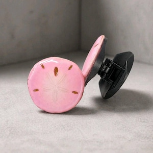 May include: A pink, round decorative object with a sand dollar design and brown accents. The object is attached to a black base. The sand dollar design features a star-like pattern.