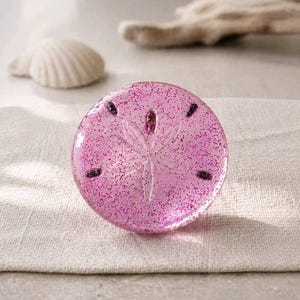 May include: A round, pink, glittery sand dollar-shaped object with a detailed starburst design in the center. The object has dark purple accents and is set on a neutral-colored linen cloth. A white seashell is in the background.
