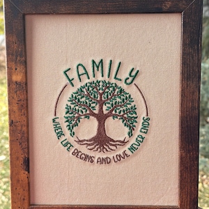 May include: Embroidered artwork in a dark wooden frame. The artwork features the word "FAMILY" above a tree design, encircled by the phrase "WHERE LIFE BEGINS AND LOVE NEVER ENDS." The embroidery uses green and brown thread on a beige fabric.