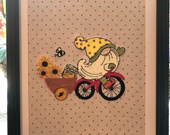 May include: Embroidered artwork featuring a gnome riding a red bicycle with a basket full of sunflowers. The background is a light beige fabric with red polka dots.