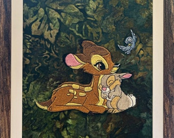 May include: Embroidered artwork featuring Bambi, Thumper, and a blue bird. The characters are set against a green and brown leaf-patterned background. The artwork is framed in a brown wooden frame with a white mat.
