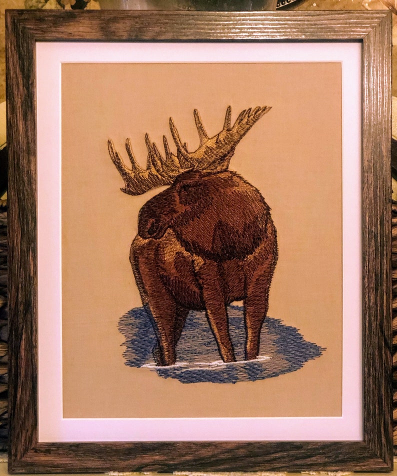 May include: Embroidered artwork of a brown moose with large antlers standing in a blue body of water. The moose is facing away from the viewer, with its back and hind legs visible.