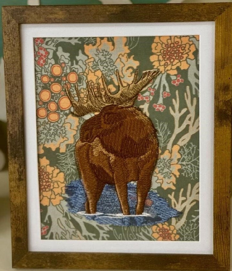 May include: Embroidered moose standing on a blue background with a floral pattern. The moose is brown and has large antlers.