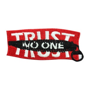 May include: A red rectangular patch with white embroidered text reading "TRUST NO ONE". The words are split by a black embroidered line. A black and white design is on the right side of the patch.