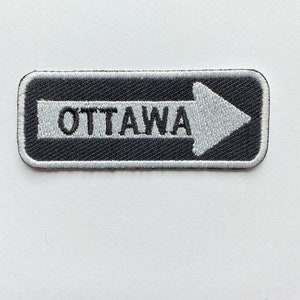 May include: A black and white embroidered patch with a white arrow pointing to the right and the word "OTTAWA" in white letters.