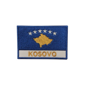 May include: A blue embroidered patch with the flag of Kosovo. The flag features a yellow outline of the country's map and six white stars in a circle. The text "KOSOVO" is embroidered in gold below the flag.