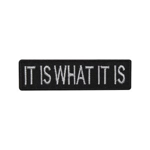 May include: A rectangular black patch with white embroidered text that reads "IT IS WHAT IT IS". The patch is a simple, bold statement, suitable for application on clothing or accessories.