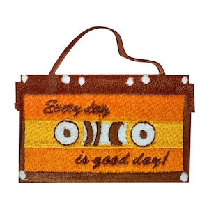 May include: An embroidered cassette tape in brown and orange with white stitching. The tape features the text "Every day is good day!" on it.