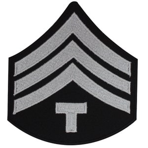 May include: A black and white embroidered patch with three silver chevrons and a silver letter "T" in the center. The patch is shaped like a chevron.