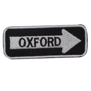 May include: Embroidered patch in black and white, featuring the word "OXFORD" and a white arrow indicating the right direction. The rectangular patch has rounded corners, designed for attachment to garments or bags.