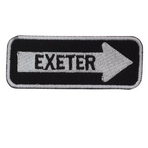 May include: A rectangular black and white embroidered patch. The patch features the word "EXETER" in white capital letters, with a white arrow pointing to the right. The patch has a black border.