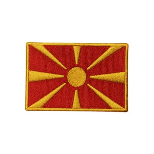May include: A red embroidered patch with a yellow sun and eight rays. The patch is a representation of the flag of North Macedonia.