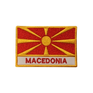 May include: An embroidered patch featuring the flag of North Macedonia. The flag is red with a yellow sun in the center and eight yellow rays extending from the sun. The patch has a white border with the word "MACEDONIA" in yellow letters.