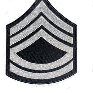 May include: A black and white embroidered patch featuring a chevron design. The patch has a dark background with three white chevrons and a curved bottom. This military-style patch is likely a rank insignia.