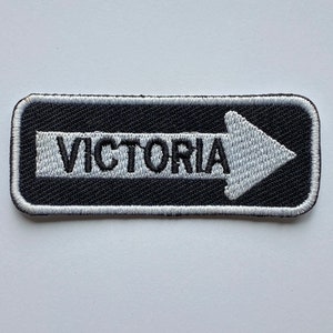 Könnte beinhalten: Weiß gestickter Patch mit schwarzem Hintergrund und einem weißen Pfeil, der nach rechts zeigt. Der Text "VICTORIA" ist in Schwarz auf dem Patch gestickt.