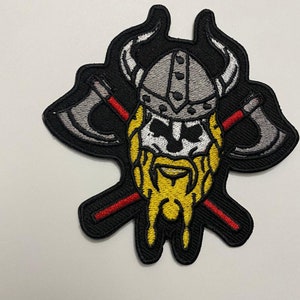 May include: Embroidered iron-on patch featuring a Viking warrior with a horned helmet, crossed axes, and a long beard. The patch is black with red and yellow accents.