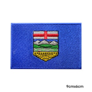 May include: An embroidered patch of the Alberta flag. The flag is blue with a coat of arms in the centre. The coat of arms features a shield with a red cross, three white mountains, a green field, and a yellow wheat field. The patch is 9 cm by 6 cm.
