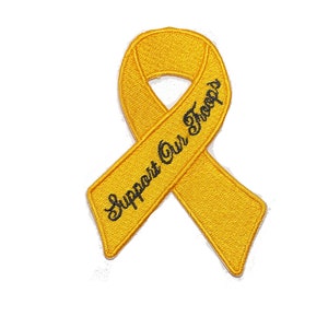 Könnte beinhalten: Ein gelber Schleifenaufnäher mit dem in schwarzer Schrift gestickten Schriftzug "Support Our Troops". Das Band ist ein Symbol der Unterstützung und des Gedenkens, das oft verwendet wird, um Solidarität zu zeigen.
