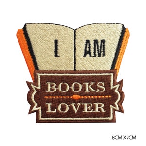 May include: Embroidered iron-on patch with a brown book design. The book has the words "I AM" on the front and "BOOKS LOVER" on a brown banner below. The patch is 8 cm by 7 cm.
