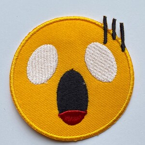 May include: Yellow embroidered iron-on patch with a surprised face with a wide open mouth and large white eyes. The patch has three black stitches on the top right corner.