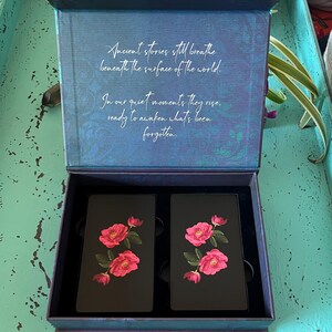 May include: An open dark blue box displaying two black cards adorned with pink rose illustrations. The box lid features the text: "Ancient stories still breathe beneath the surface of the world."
