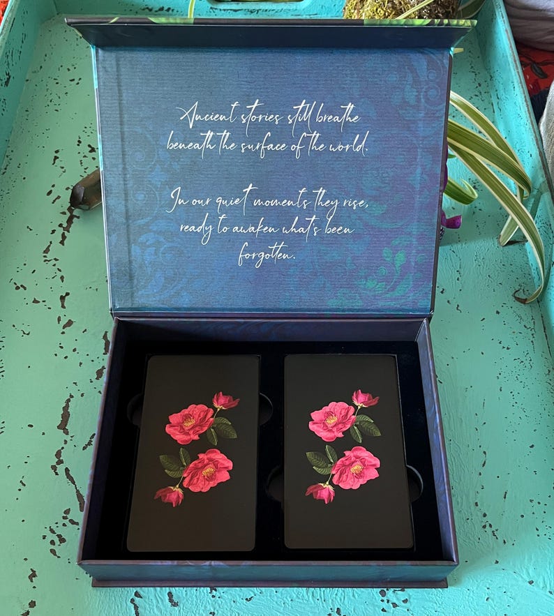 May include: An open dark blue box displaying two black cards adorned with pink rose illustrations. The box lid features the text: "Ancient stories still breathe beneath the surface of the world."