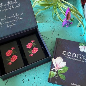 May include: A tarot card set and book. The cards are black with pink rose illustrations. The book cover is dark blue with a white flower and the title "CODEX The Myth Weaver Tarot". A purple rabbit figurine is also visible.