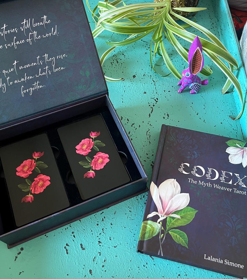 May include: A tarot card set and book. The cards are black with pink rose illustrations. The book cover is dark blue with a white flower and the title "CODEX The Myth Weaver Tarot". A purple rabbit figurine is also visible.