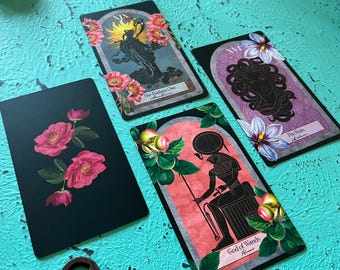 Könnte beinhalten: Eine Sammlung von Tarotkarten mit floralen und mythologischen Illustrationen. Die Karten zeigen Bilder einer strahlenden Figur, eines Medusa-ähnlichen Gesichts und einer ägyptischen Gottheit. Ein antiker Schlüssel liegt in der Nähe der Karten auf einer türkisfarbenen Oberfläche.