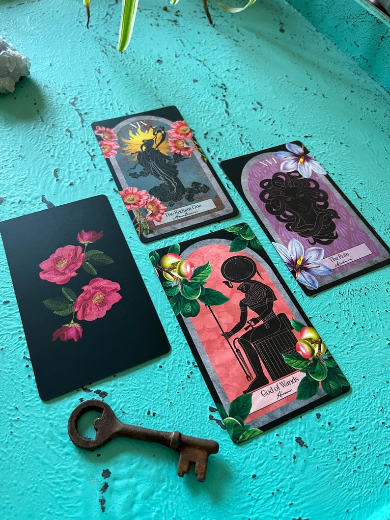 May include: A selection of tarot cards with floral and mythological imagery. The cards depict a radiant figure, a Medusa-like face, and an Egyptian deity. A vintage key is placed near the cards on a turquoise surface.