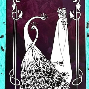 May include: A tarot card featuring a white peacock with a crown facing a robed figure, set against a dark purple background. The card is framed with white decorative elements and the text "QUEEN OF WANDS" at the bottom.
