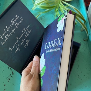 May include: A book titled "CODEX The Myth Weaver Tarot" with a floral design on the cover. The book is held open, revealing text inside the cover. The book is in a dark blue box, with the author's name, "Lalania Simone", visible.