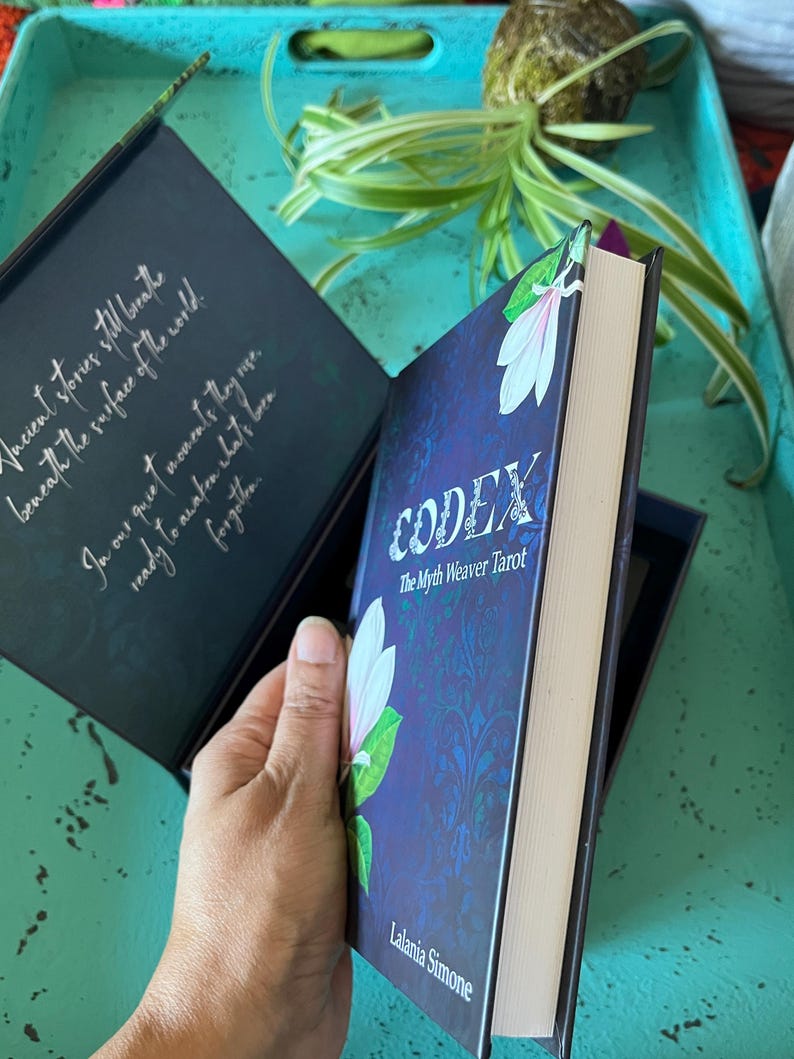 May include: A book titled "CODEX The Myth Weaver Tarot" with a floral design on the cover. The book is held open, revealing text inside the cover. The book is in a dark blue box, with the author's name, "Lalania Simone", visible.