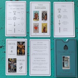 May include: A tarot card deck with a teal box and several cards laid out. The cards feature illustrations and text, including suits, elements, and card meanings. The box reads "COSMIC SEED sacred goods" and "Tarot Flashcards".