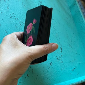 May include: A deck of tarot cards featuring pink rose illustrations on a dark teal background. The card edges are black. The deck is held in a hand against a turquoise surface.