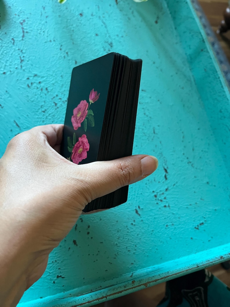May include: A deck of tarot cards featuring pink rose illustrations on a dark teal background. The card edges are black. The deck is held in a hand against a turquoise surface.