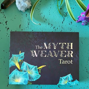 May include: A tarot card deck titled "The Myth Weaver Tarot" with a dark blue background and turquoise morning glory flowers. The text is in white and the deck is on a teal surface.