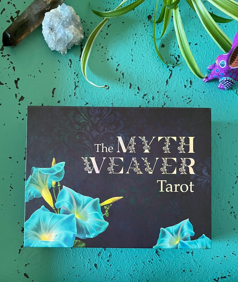 May include: A tarot card deck titled "The Myth Weaver Tarot" with a dark blue background and turquoise morning glory flowers. The text is in white and the deck is on a teal surface.