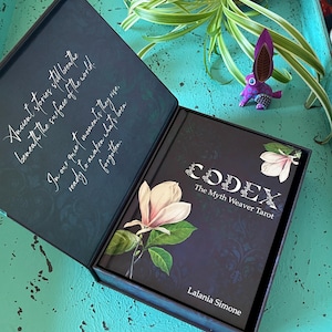 May include: A dark blue box set containing a tarot deck titled "CODEX The Myth Weaver Tarot" by Lalania Simone. The box and deck feature floral illustrations and text. A small purple rabbit figurine is visible.