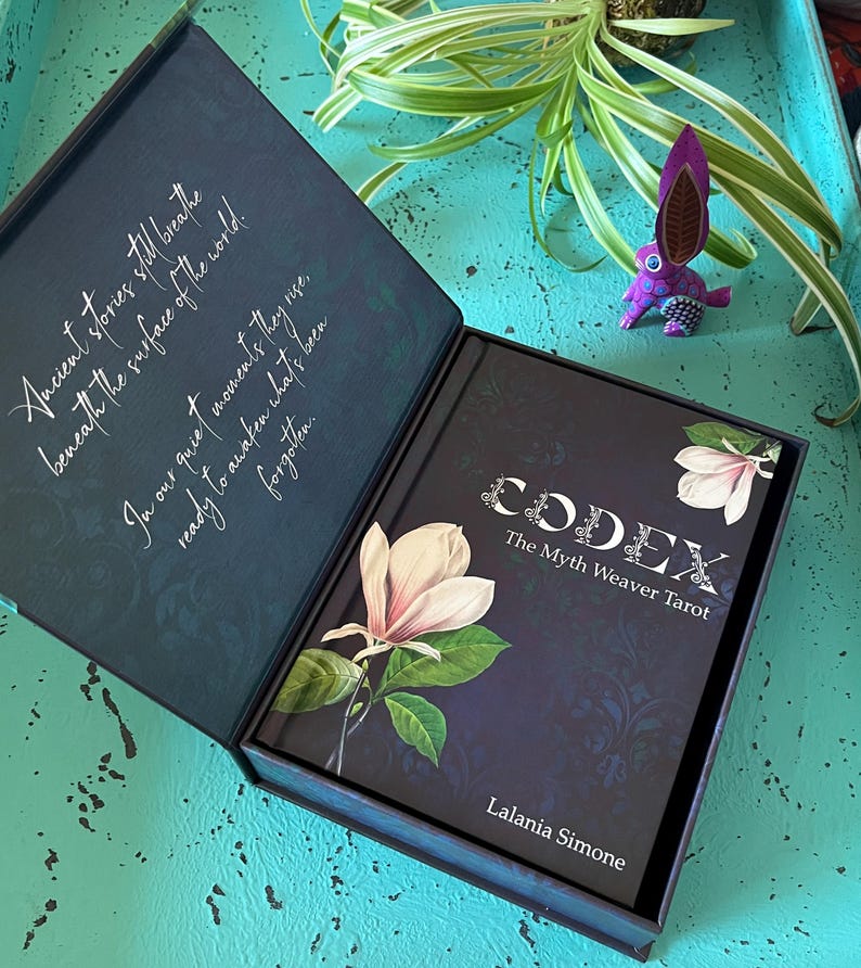 May include: A dark blue box set containing a tarot deck titled "CODEX The Myth Weaver Tarot" by Lalania Simone. The box and deck feature floral illustrations and text. A small purple rabbit figurine is visible.