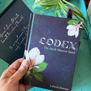May include: A hardback book titled "CODEX The Myth Weaver Tarot" with a dark blue cover featuring a floral design and a white magnolia. The author's name, Lalania Simone, is at the bottom. Another book with text is in the background.